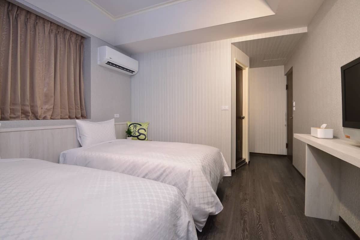 Habitación de hotel con dos camas, un televisor de pantalla plana, un equipo de aire acondicionado y una mesita de noche pequeña con una caja de pañuelos.