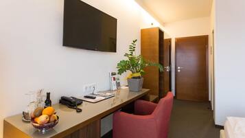 A hotel room with a desk, a television mounted on the wall, a potted plant, a red chair, and a bed.