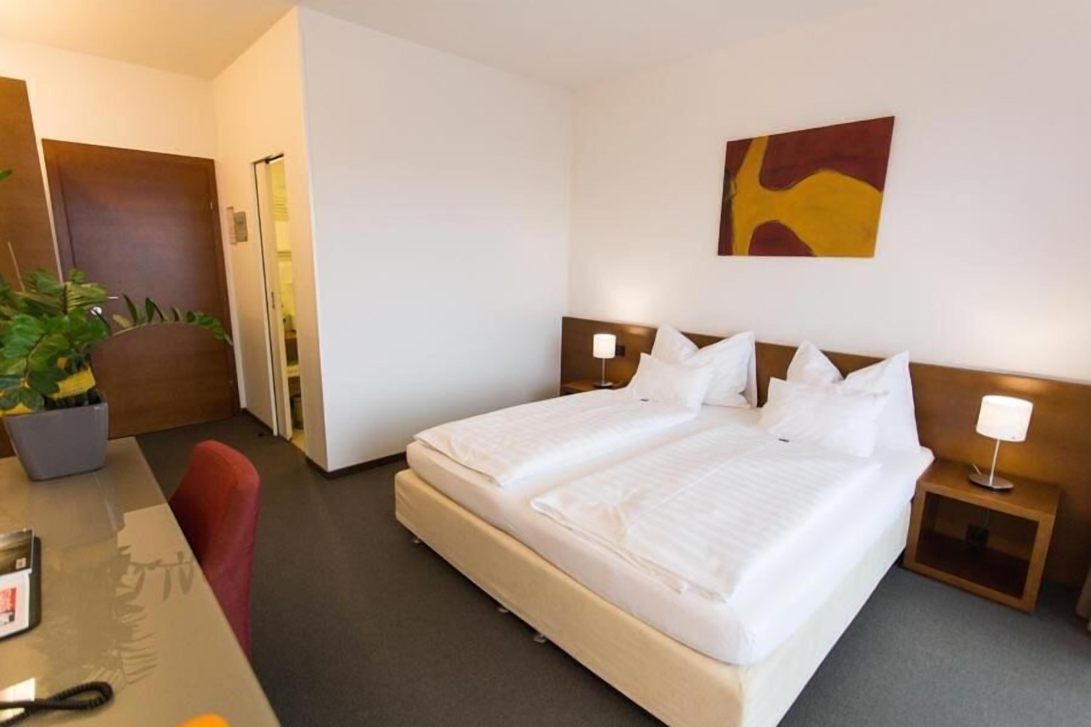 A hotel room with a large bed, a desk with a chair, a potted plant, and a painting on the wall.