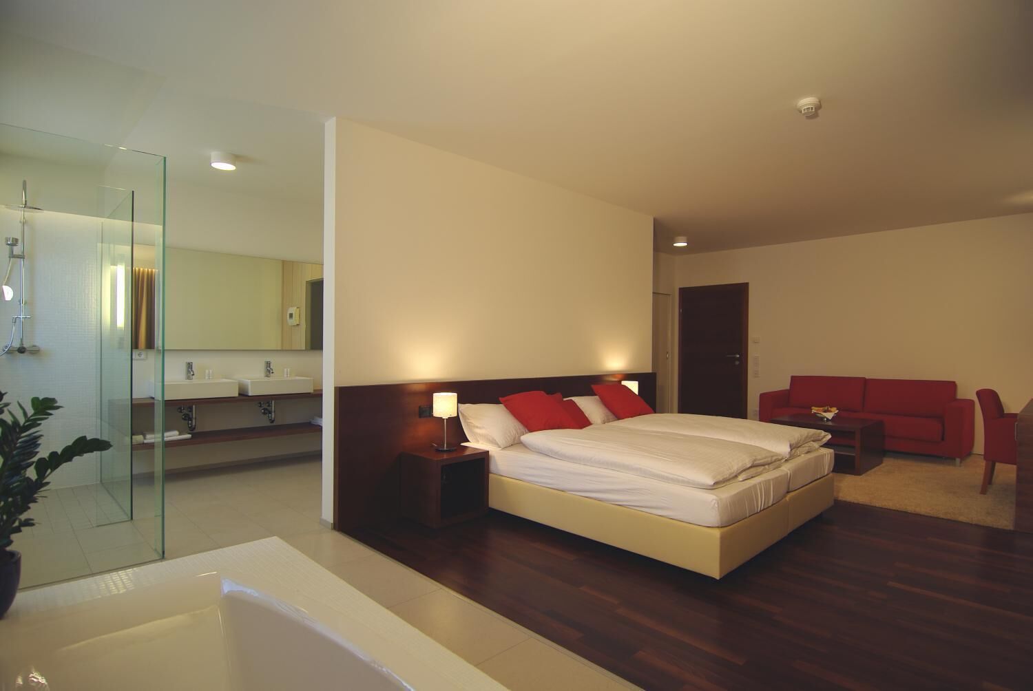 Een moderne hotelkamer met een groot bed, een douchecabine met glazen wanden, een rode bank en een kleine tafel.