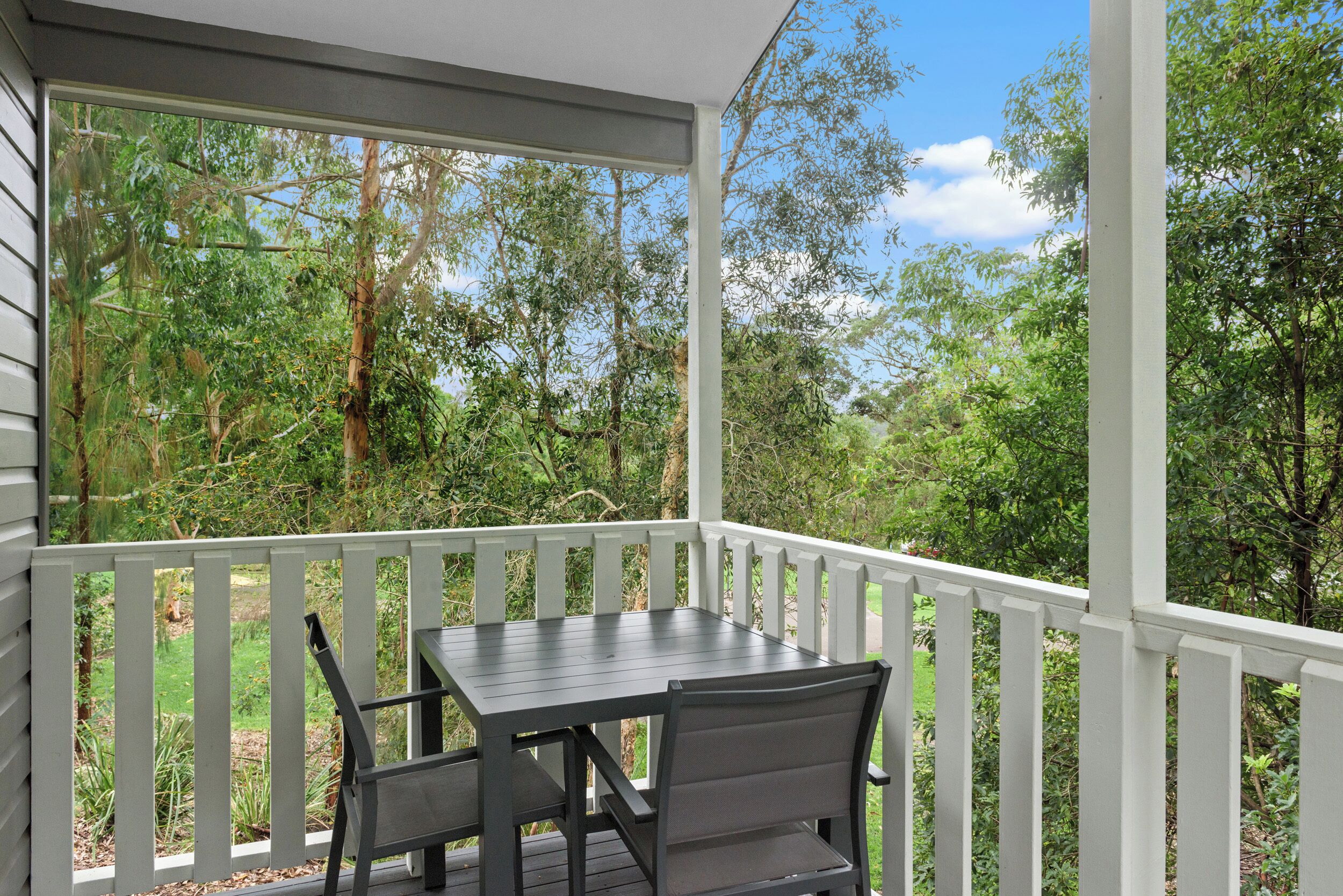 Un patio cubierto al aire libre con una mesa y sillas, rodeado por una barandilla blanca, con vistas a un exuberante jardín y árboles.