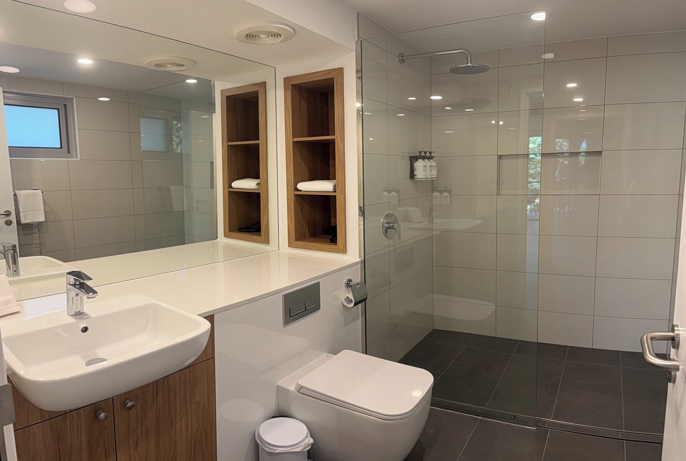 Un baño moderno con lavamanos, espejo, armarios de madera, inodoro y cabina de ducha con mampara de vidrio.