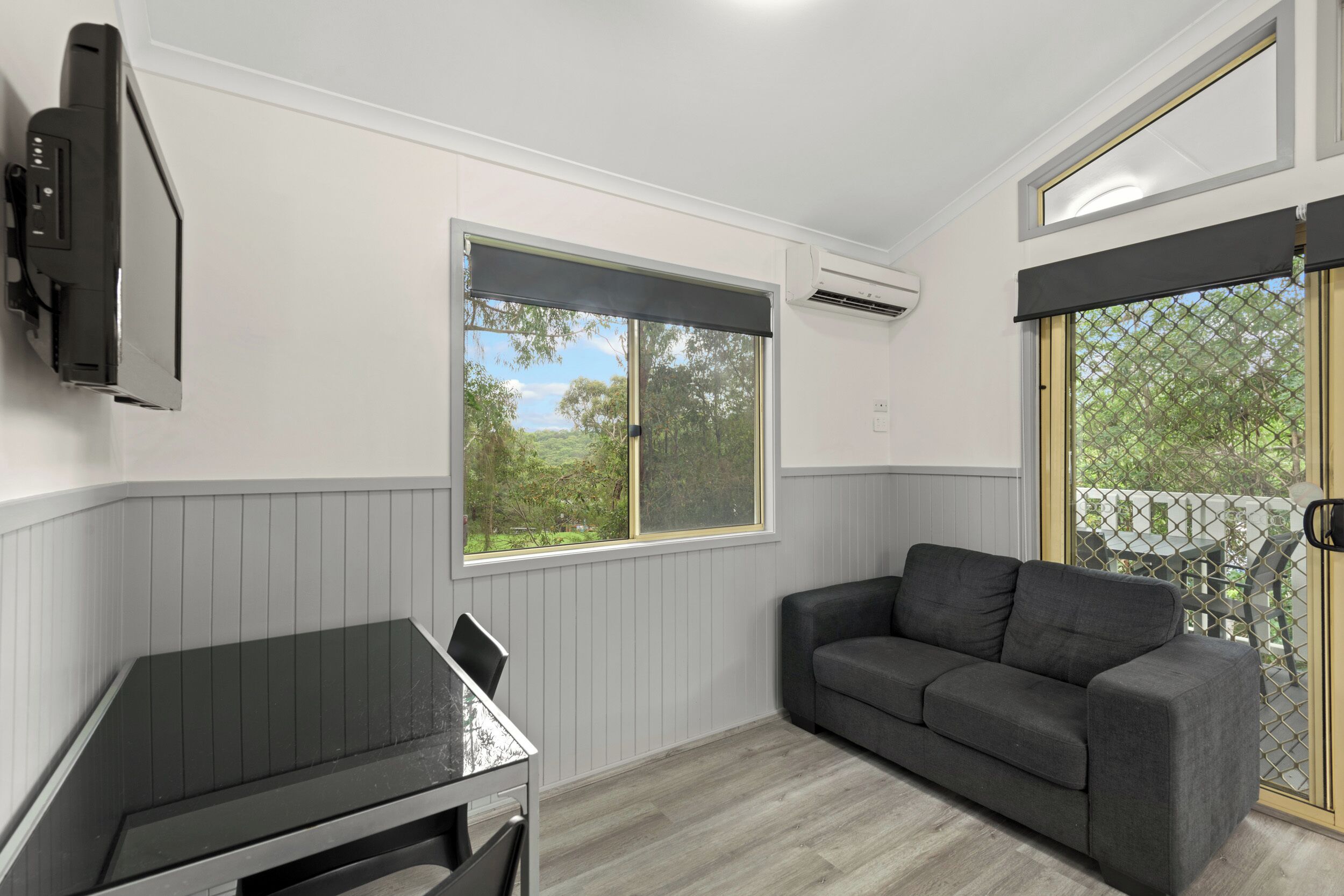 A room with a grey couch, a small table, a window with a view of greenery, and an air conditioning unit.