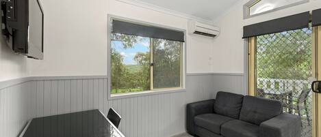 A room with a grey couch, a small table, a window with a view of greenery, and an air conditioning unit.