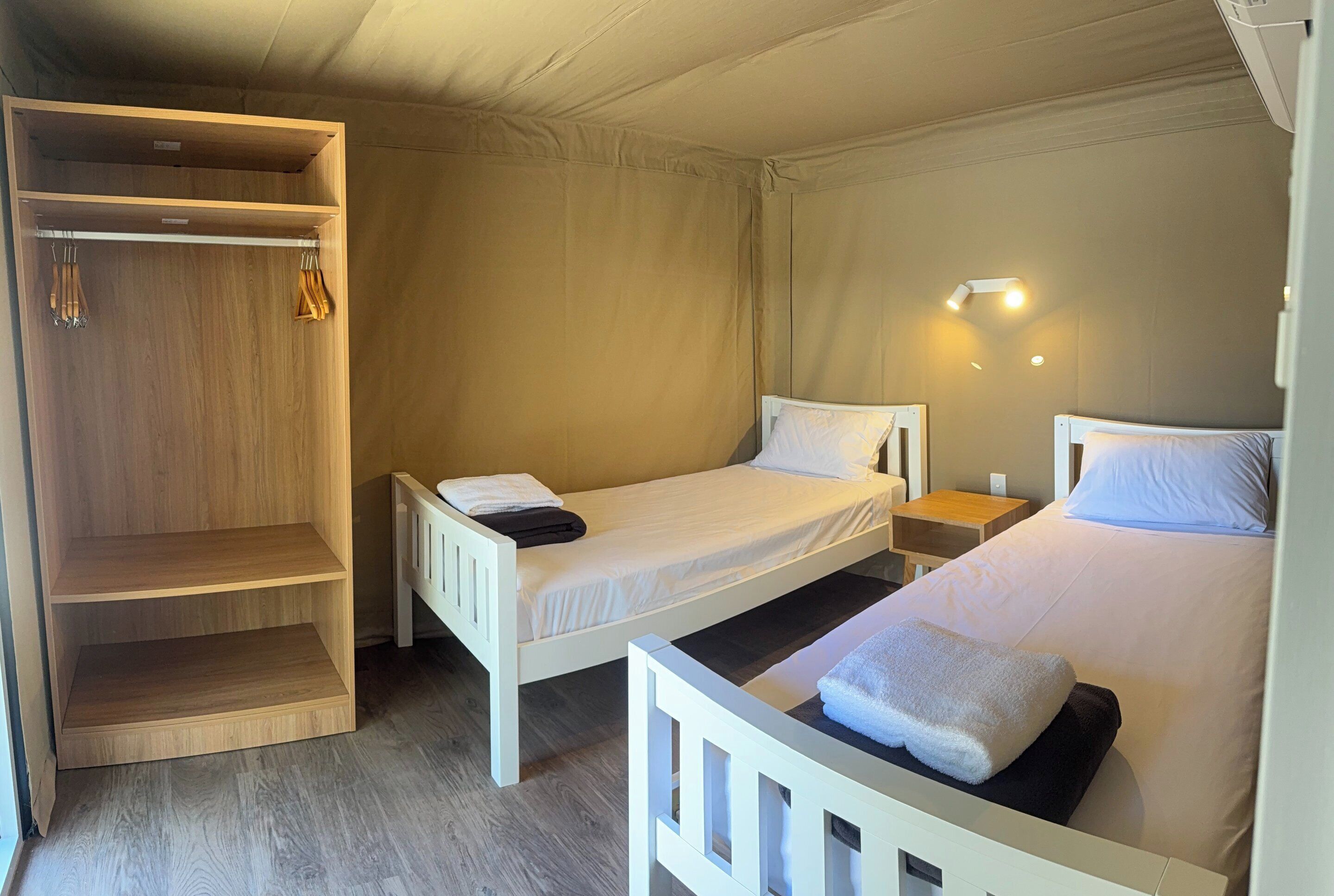 A single bedroom with two beds, a wooden wardrobe, and a bedside table with a lamp.