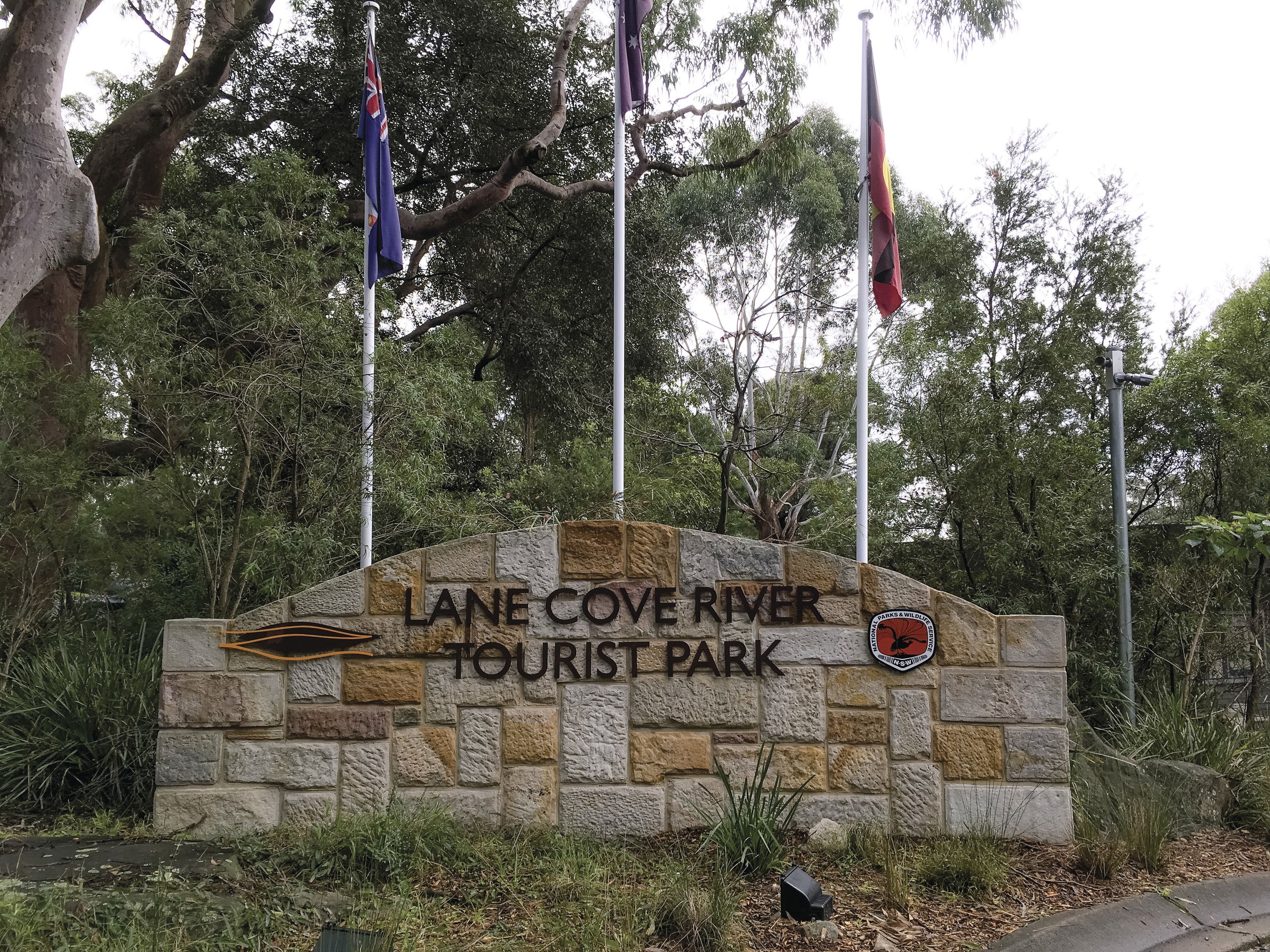 A stone sign with "LANE COVE RIVER TOURIST PARK" and a logo for "PARKS & WILDLIFE" on a stone wall.