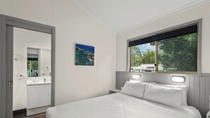 Quarto com uma cama grande, janela com vista para o verde, banheiro com espelho e um quadro na parede.