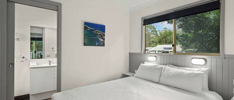 A bedroom with a large bed, a window with a view of greenery, a bathroom with a mirror, and a painting on the wall.