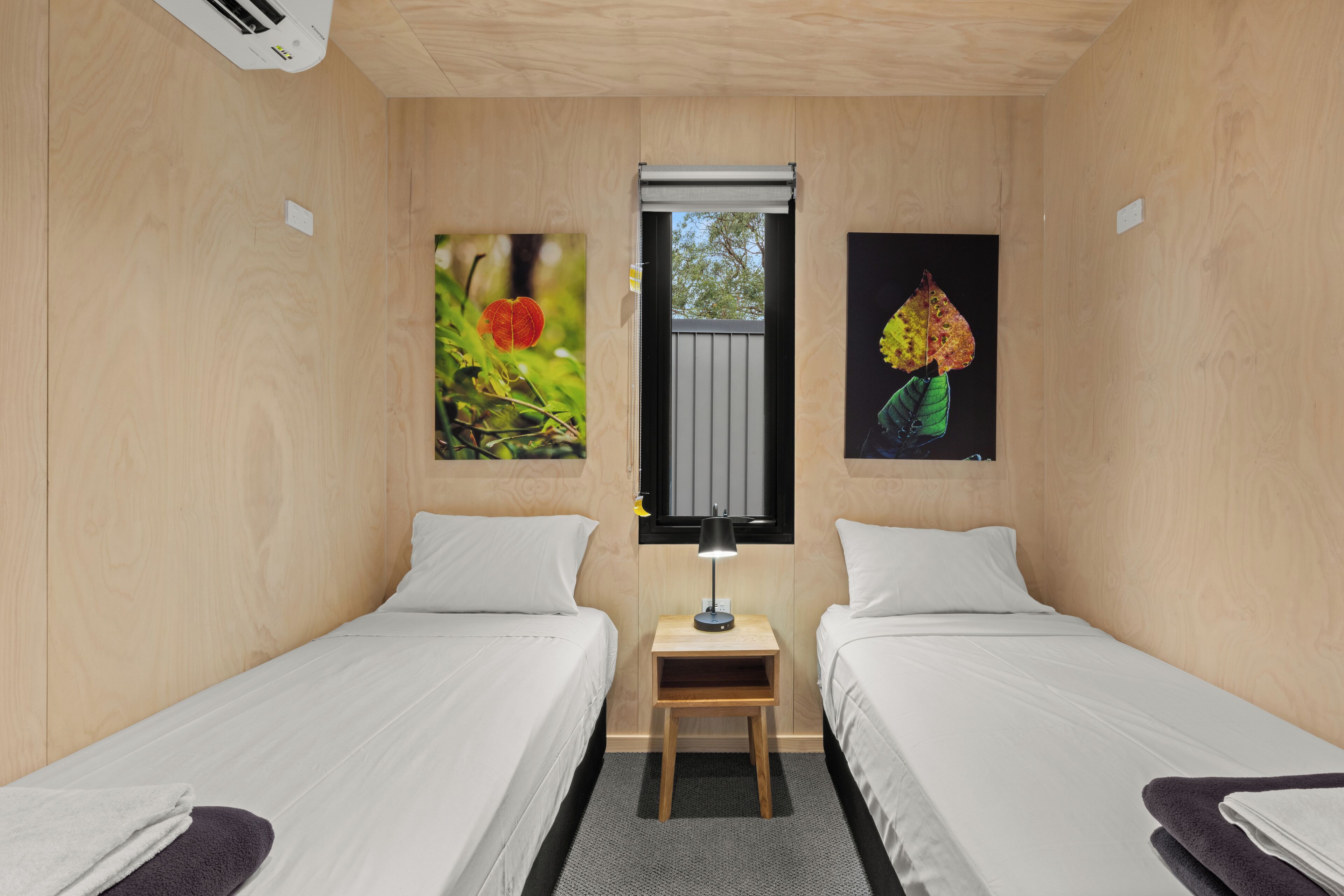Quarto com duas camas, uma mesinha com abajur, e duas fotos emolduradas nas paredes.