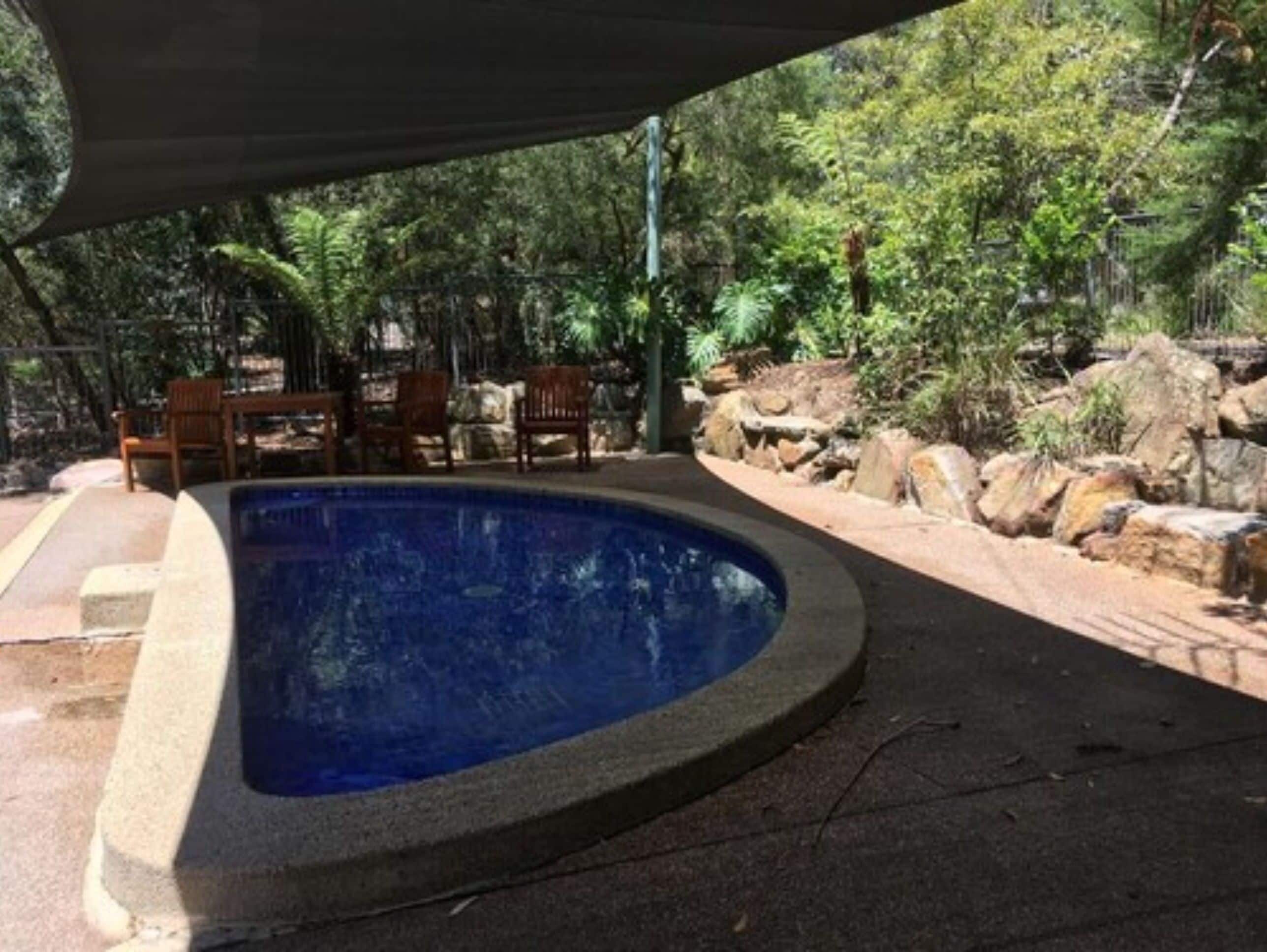 Une piscine à bord incurvé, entourée d’une bordure en pierre et agrémentée d’un espace ombragé avec des chaises en bois.