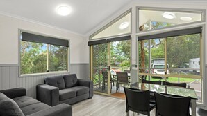 Quarto moderno com sofás cinzas, mesa de vidro e cadeiras, janelas amplas com persianas e vista para o exterior.