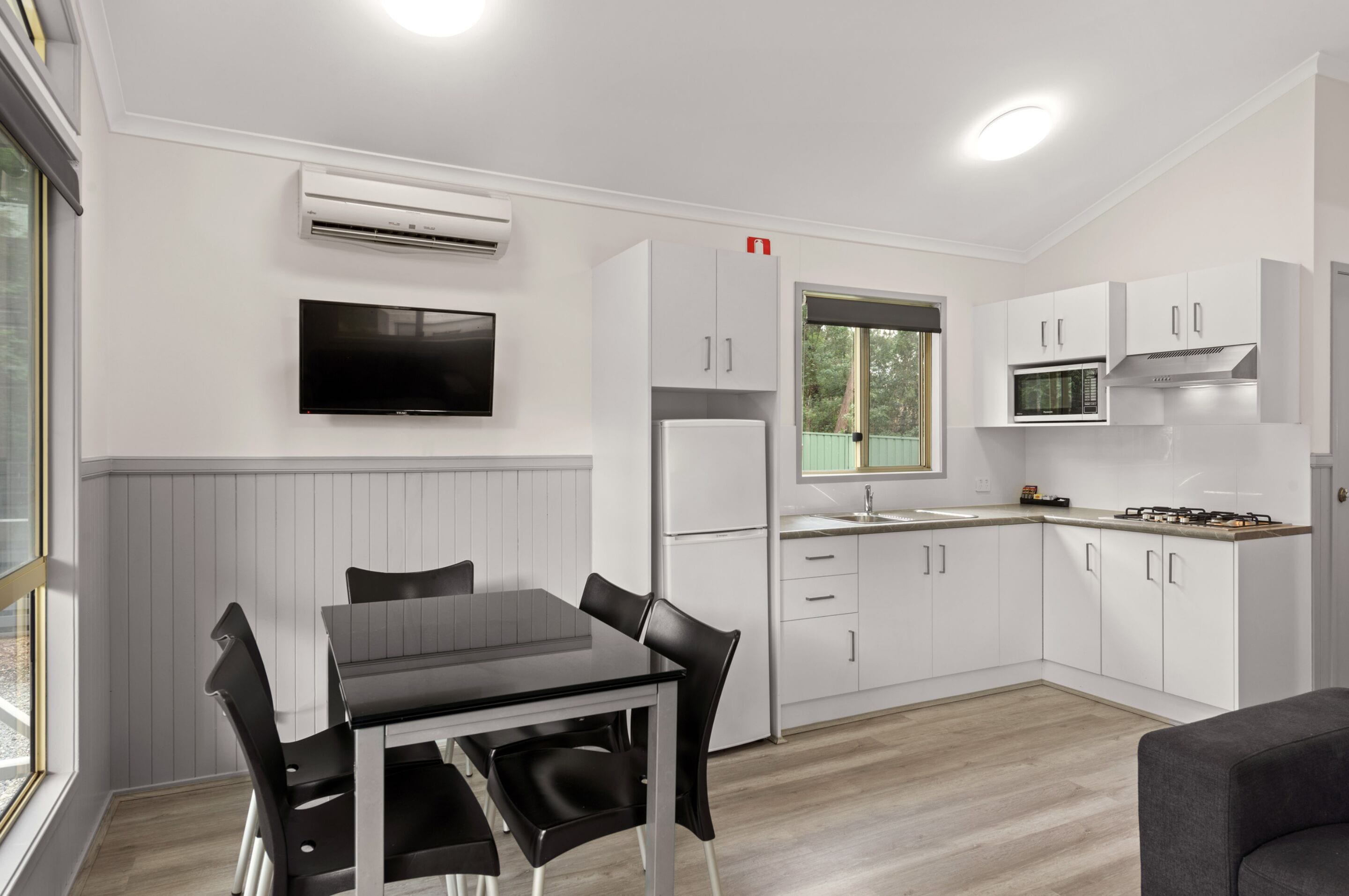 Une cuisine moderne avec des armoires blanches, un four à micro-ondes encastré et une cuisinière à gaz. On y trouve une table à manger avec des chaises, ainsi qu’un téléviseur à écran plat fixé au mur.