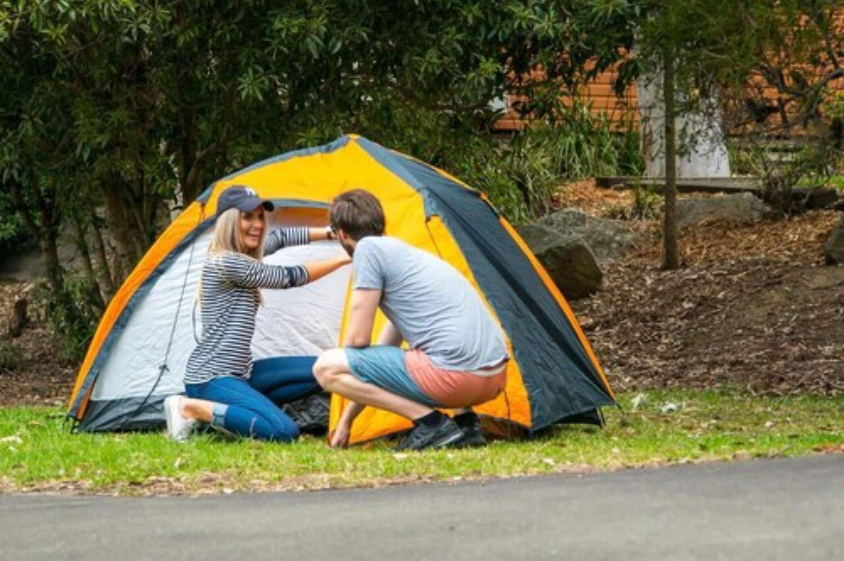 Two people setting up a tent in a grassy area with trees and a brick wall in the background.