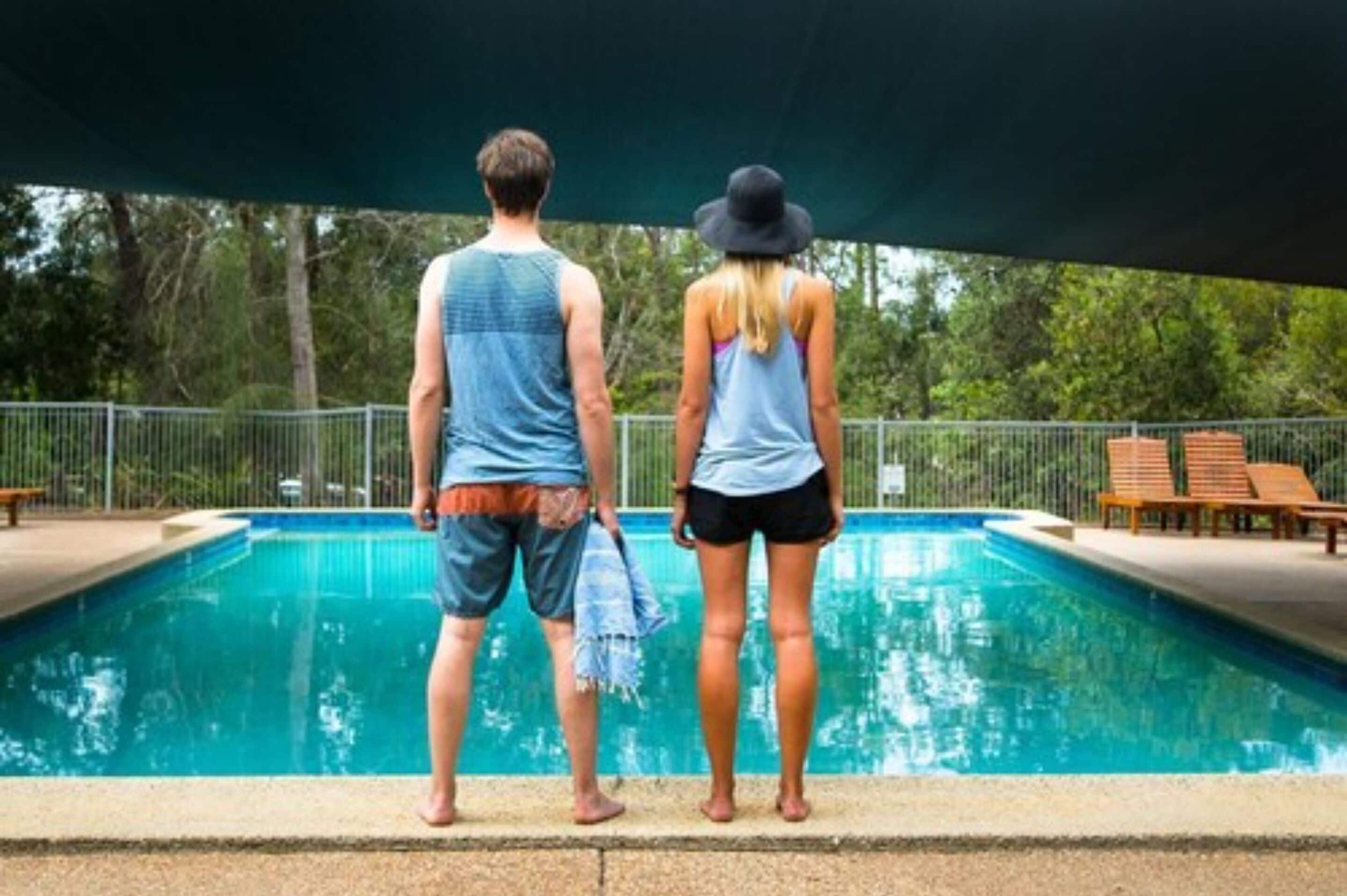 A swimming pool with two people standing at the edge, a shaded area, and a fence in the background.