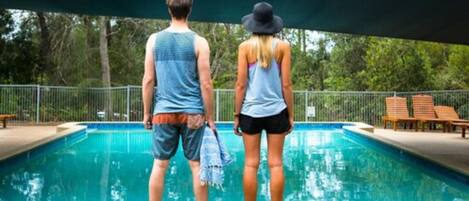 A swimming pool with two people standing at the edge, a shaded area, and a fence in the background.