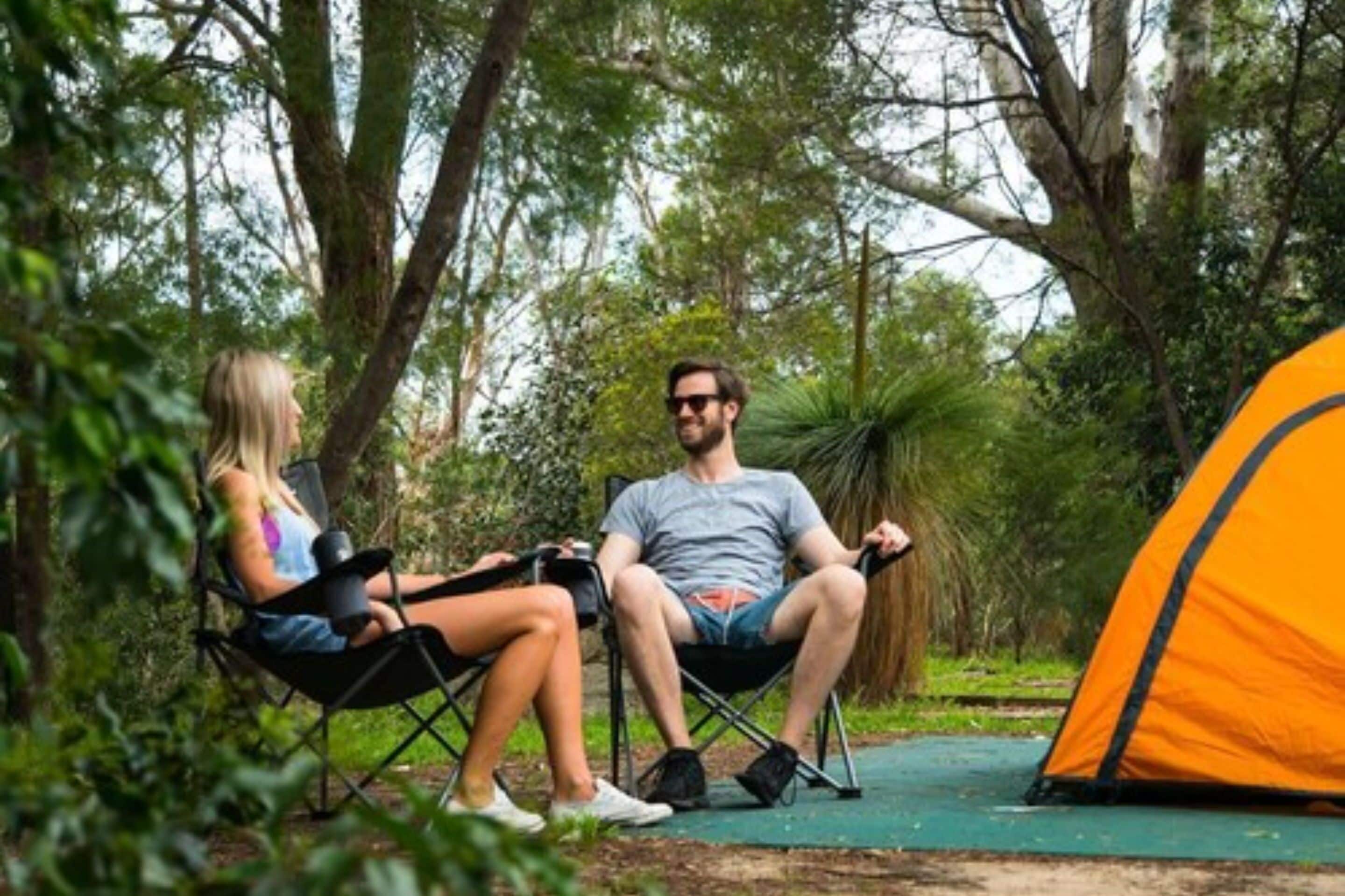 Two people sitting on camping chairs in a forested area with a tent nearby.