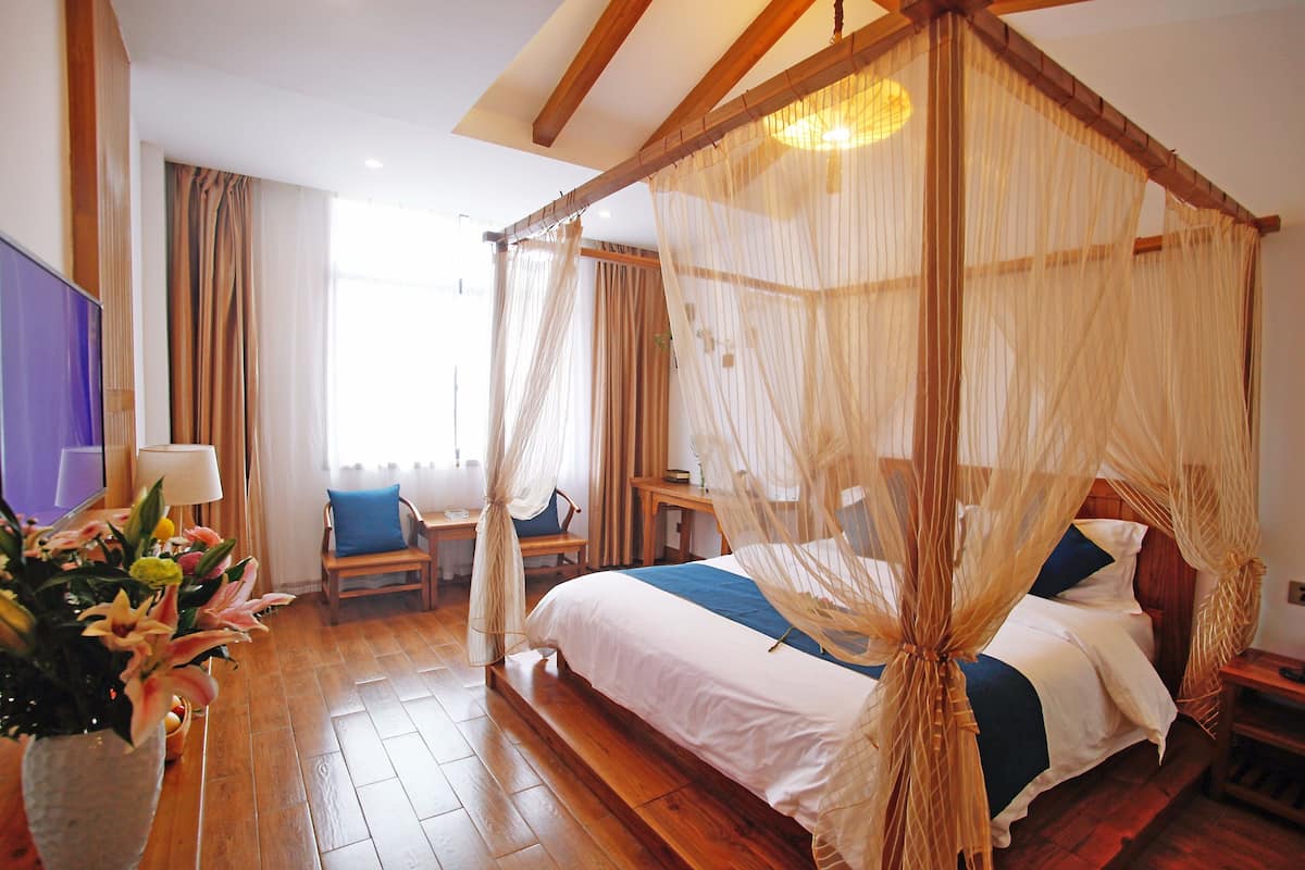 Un lit à baldaquin à quatre poteaux, avec une tête de lit en bois, un plancher en bois, une table de chevet avec une lampe et une chaise avec un coussin.