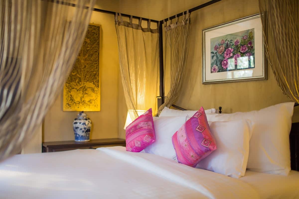 A bed with white linens and pink pillows, a framed floral artwork on the wall, and a vase on the nightstand.