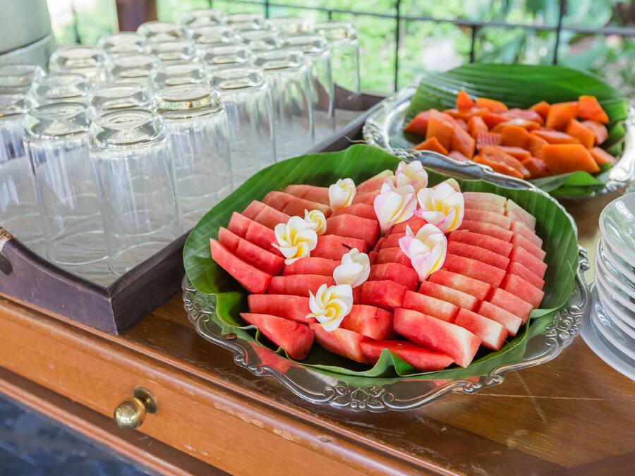Un tavolo per buffet con fette di anguria su una foglia di banano, una ciotola di carote a rondelle e bicchieri di vetro.