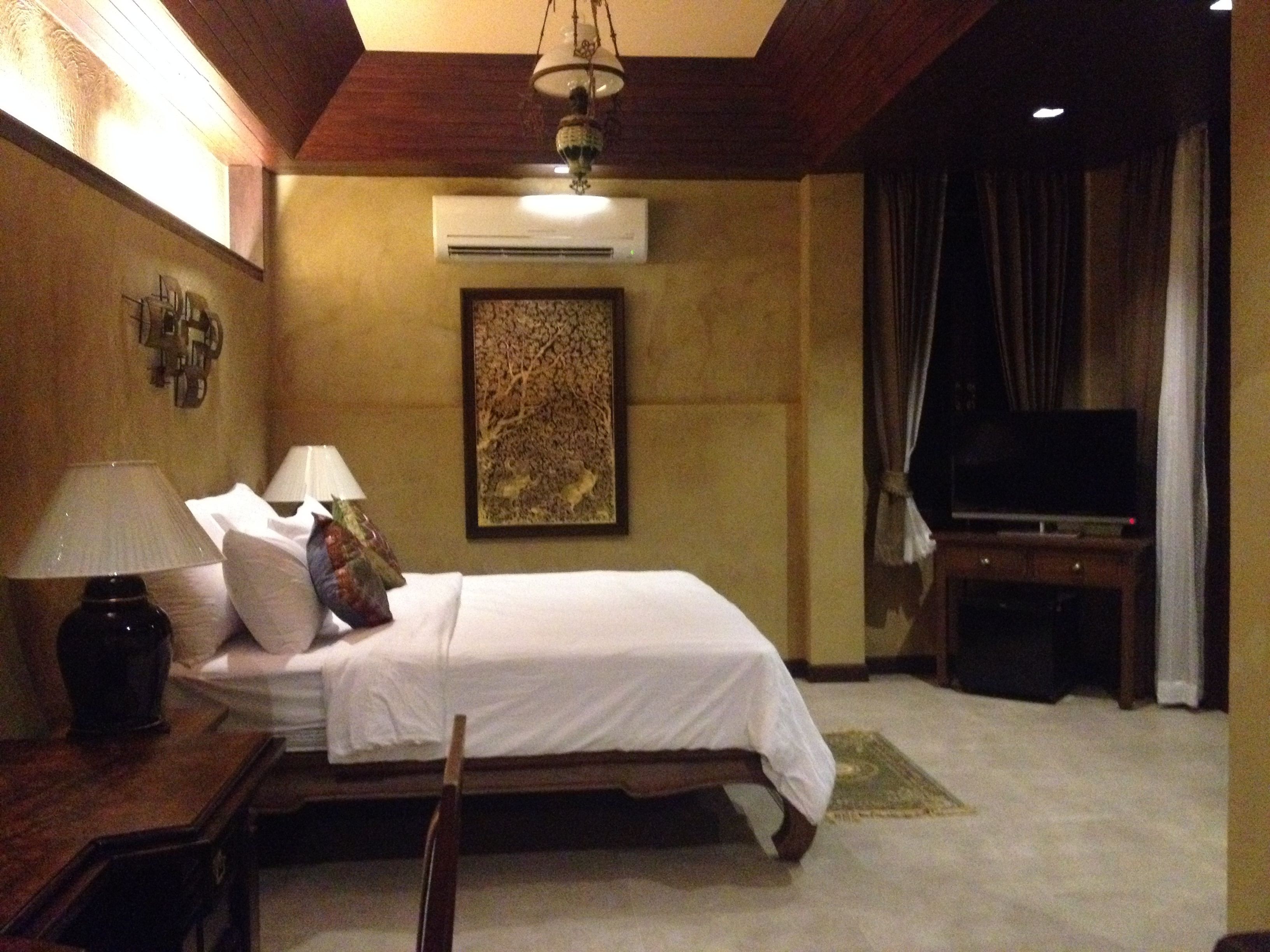 A hotel room with a bed, two bedside lamps, an air conditioner, a television on a stand, and a framed artwork on the wall.