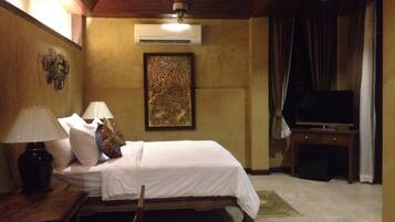 A hotel room with a bed, two bedside lamps, an air conditioner, a television on a stand, and a framed artwork on the wall.