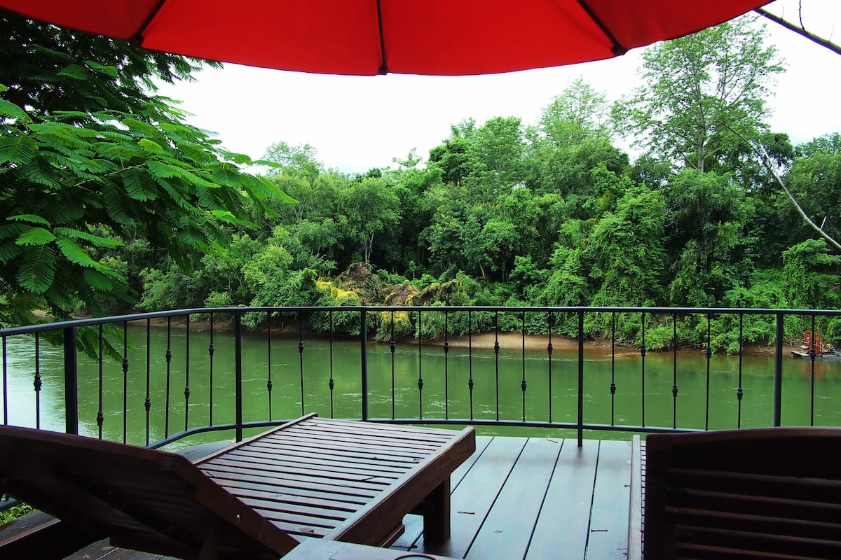 A wooden deck with a red umbrella, overlooking a river with dense greenery and a metal railing.