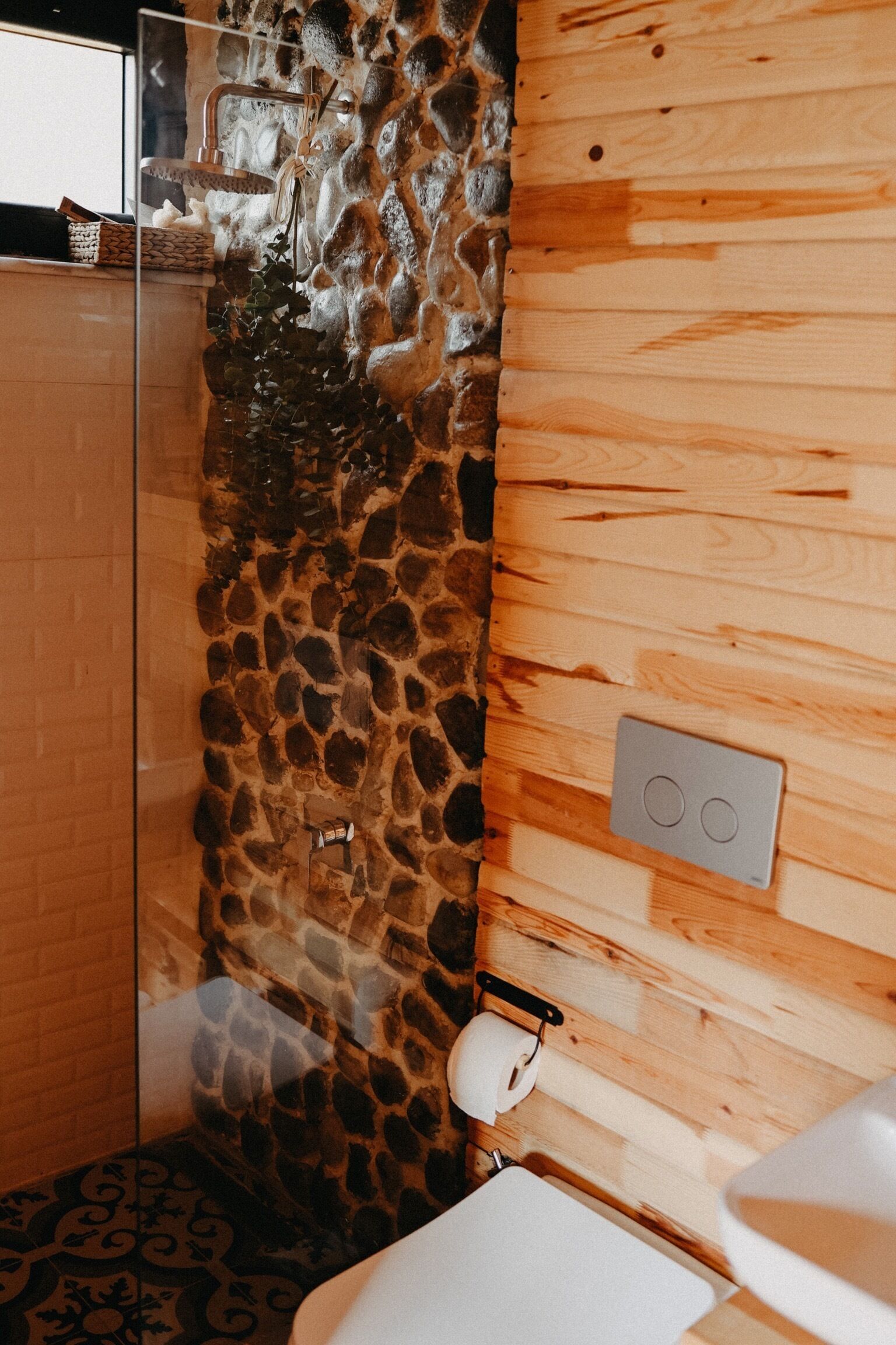 Un baño con pared de piedra, paneles de madera, cabina de ducha de vidrio, lavabo blanco y dispensador de toallas.