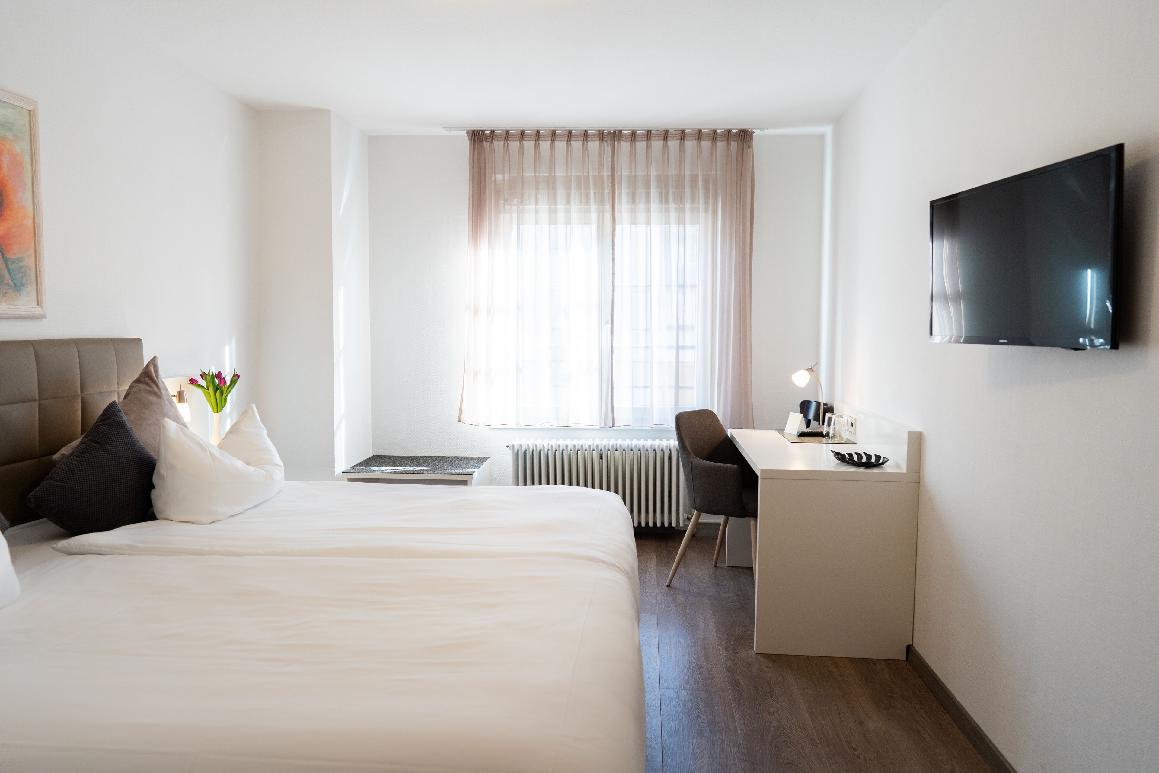 A hotel room with a bed, a desk, a chair, a radiator, and a TV mounted on the wall.