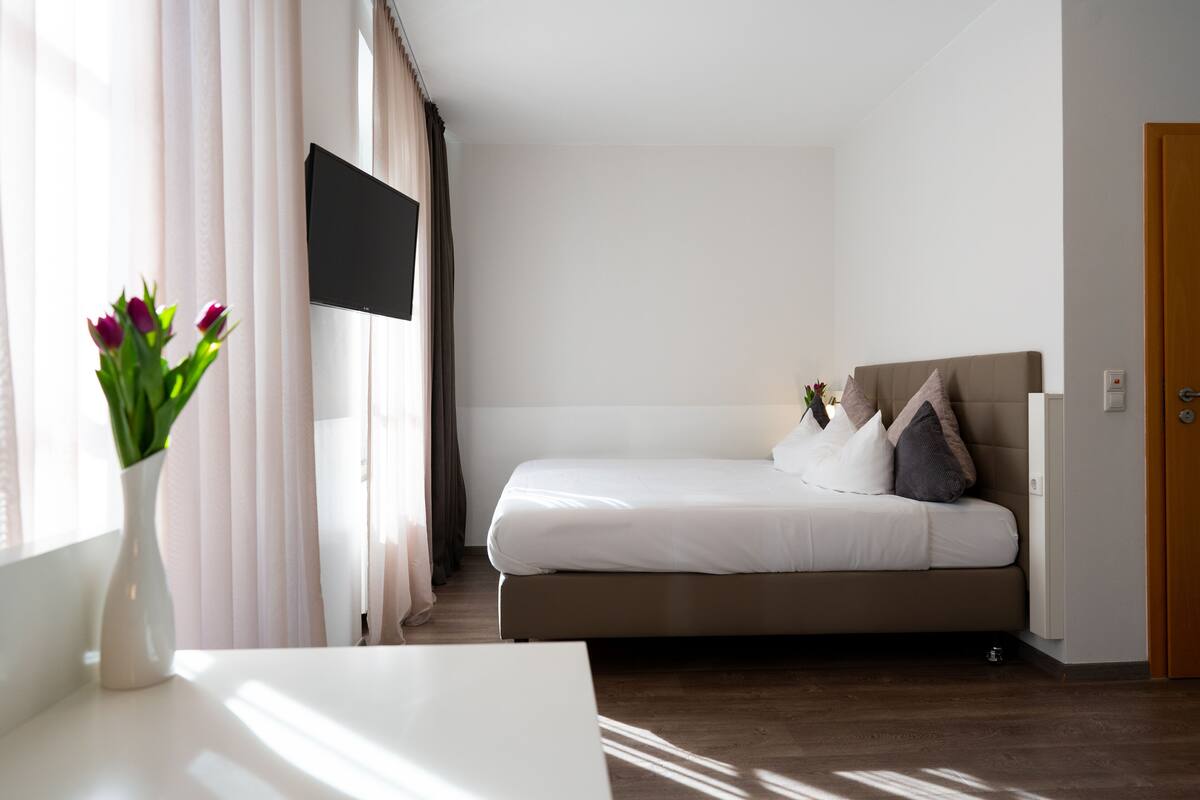 A bedroom with a bed, a TV mounted on the wall, a vase with flowers, and a wooden door.