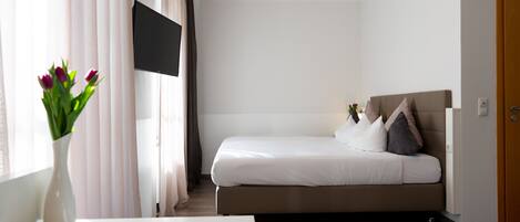 Een slaapkamer met een bed, een aan de muur bevestigde televisie, een vaas met bloemen en een houten deur.