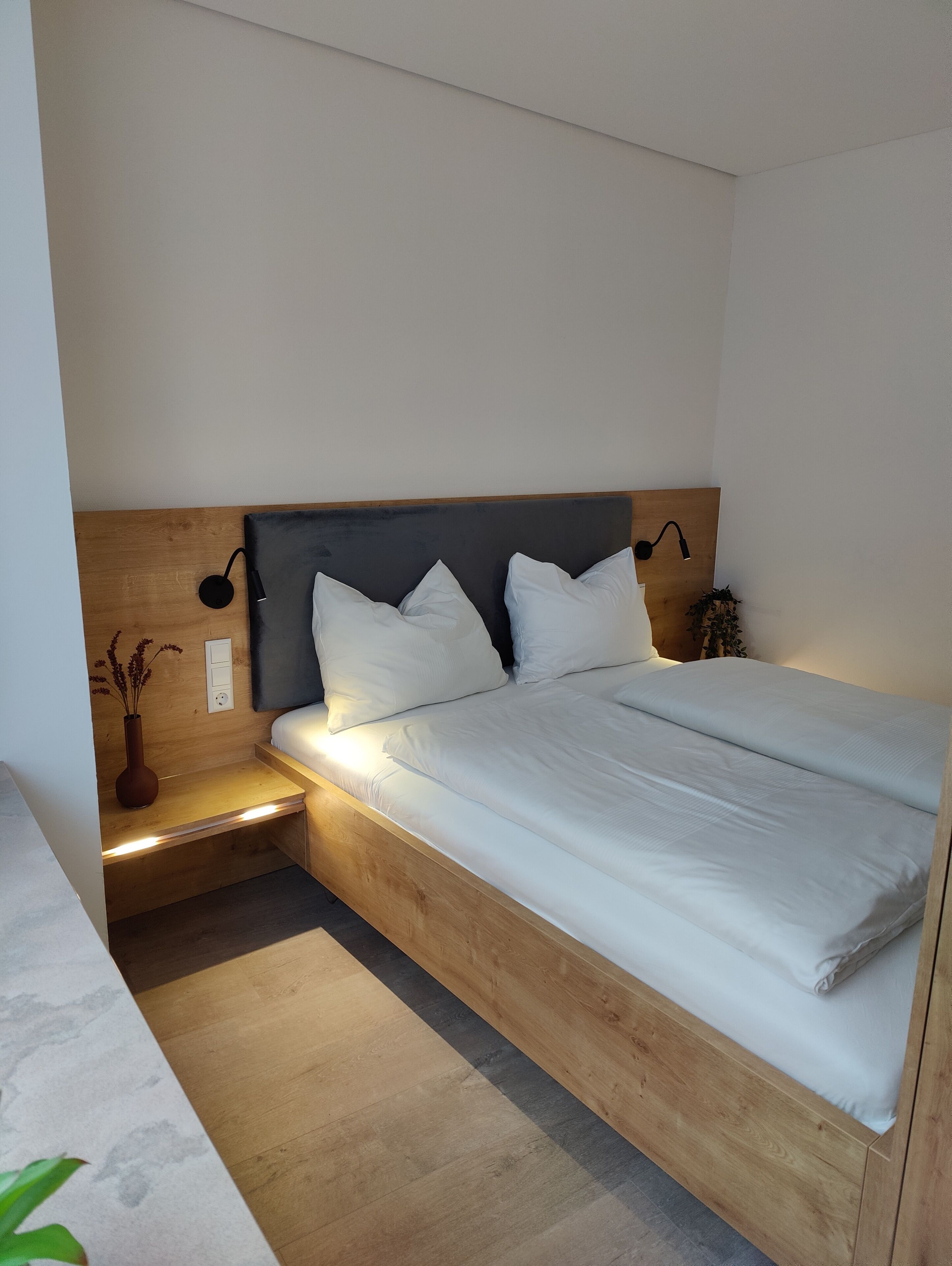 Une chambre à coucher moderne avec une tête de lit en bois, une table de chevet ornée d’un vase et une petite table de nuit.