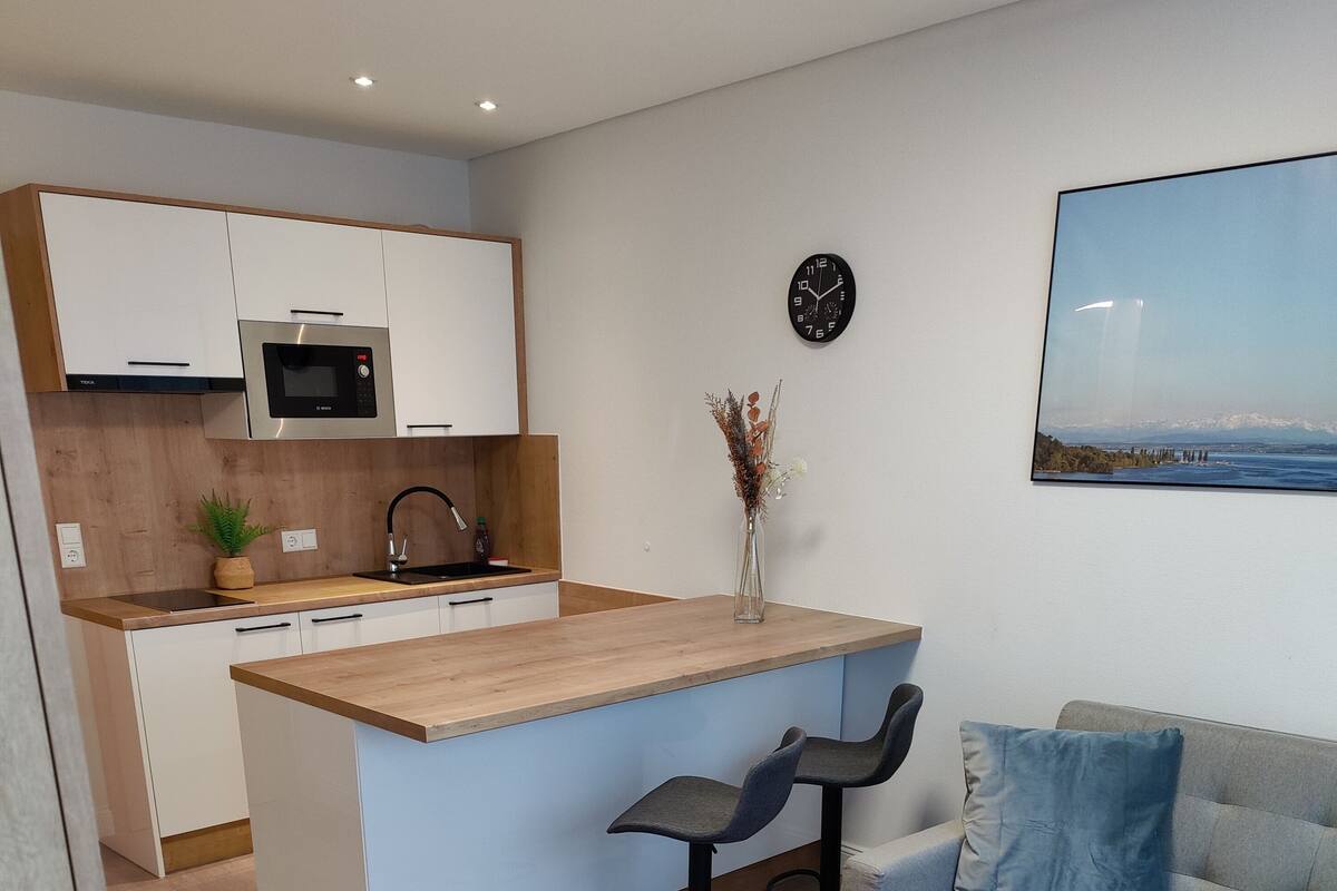 A modern kitchen with white cabinets, a wooden countertop, and a built-in microwave. There is a dining area with a glass table and two chairs, and a gray sofa with blue cushions.