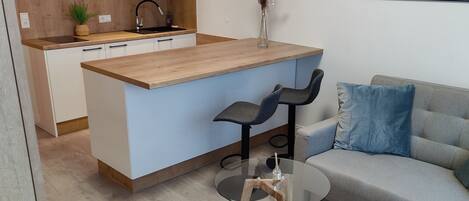 A modern kitchen with white cabinets, a wooden countertop, and a built-in microwave. There is a dining area with a glass table and two chairs, and a gray sofa with blue cushions.