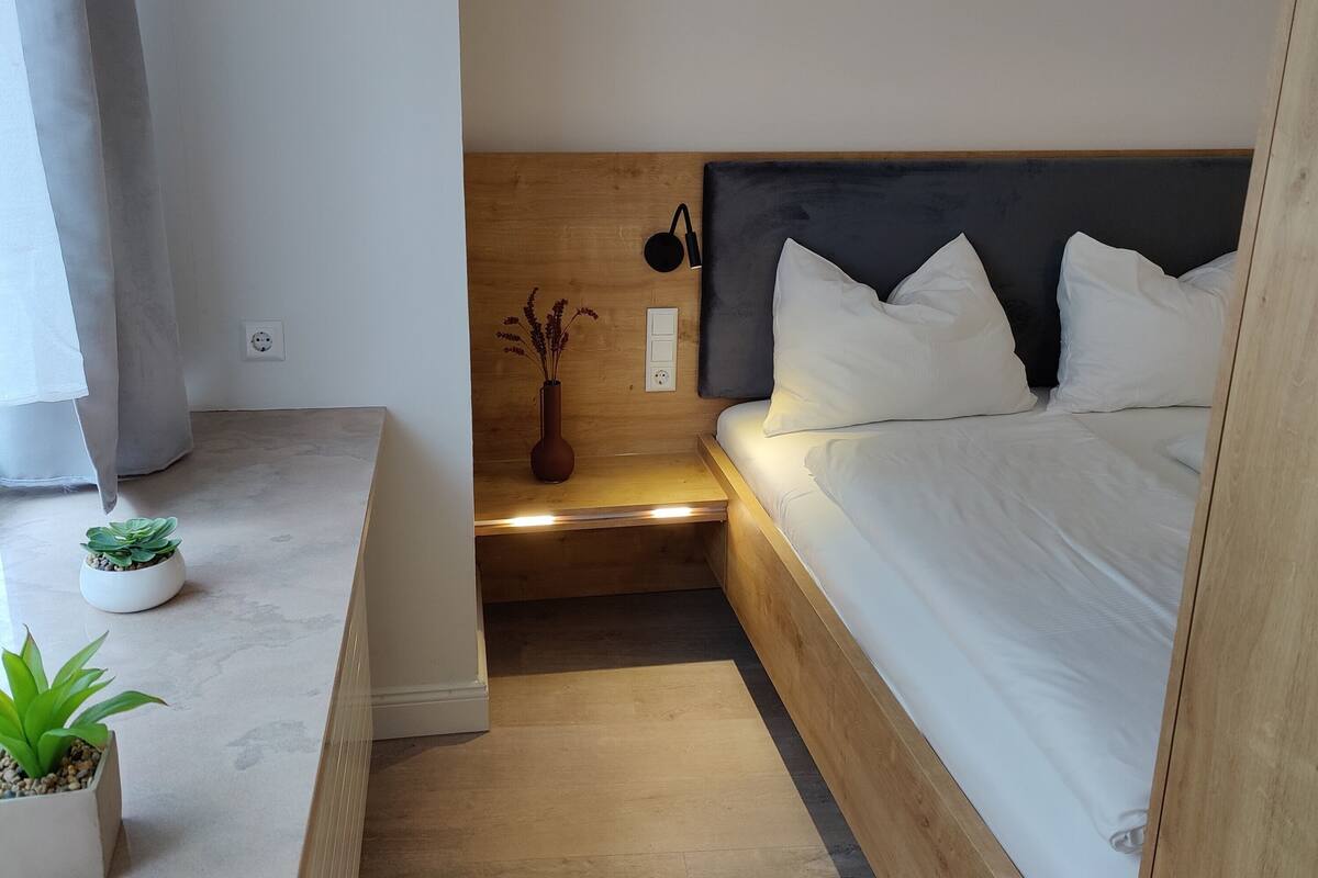 A modern bedroom with a wooden headboard, a bedside table with a vase, and a small potted plant.