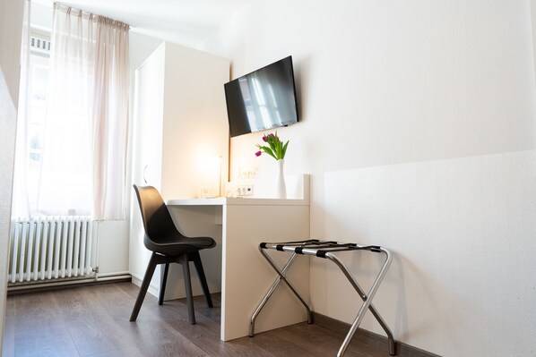 Una habitación con escritorio, silla, televisor montado en la pared, radiador y ventana con cortinas.