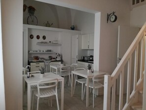 A compact kitchen with white cabinets, a dining area with white chairs, and a staircase. - InCentro b&b (Porto Cesareo)