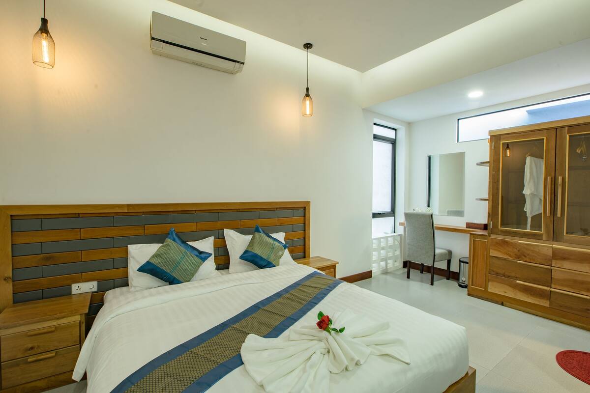 A hotel room with a wooden headboard, a bed with white bedding and blue accents, a wooden wardrobe, and a small desk with a chair.