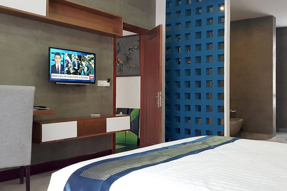 A modern hotel room with a bed, a TV mounted on the wall, a desk with a chair, and a bathroom with a large window.
