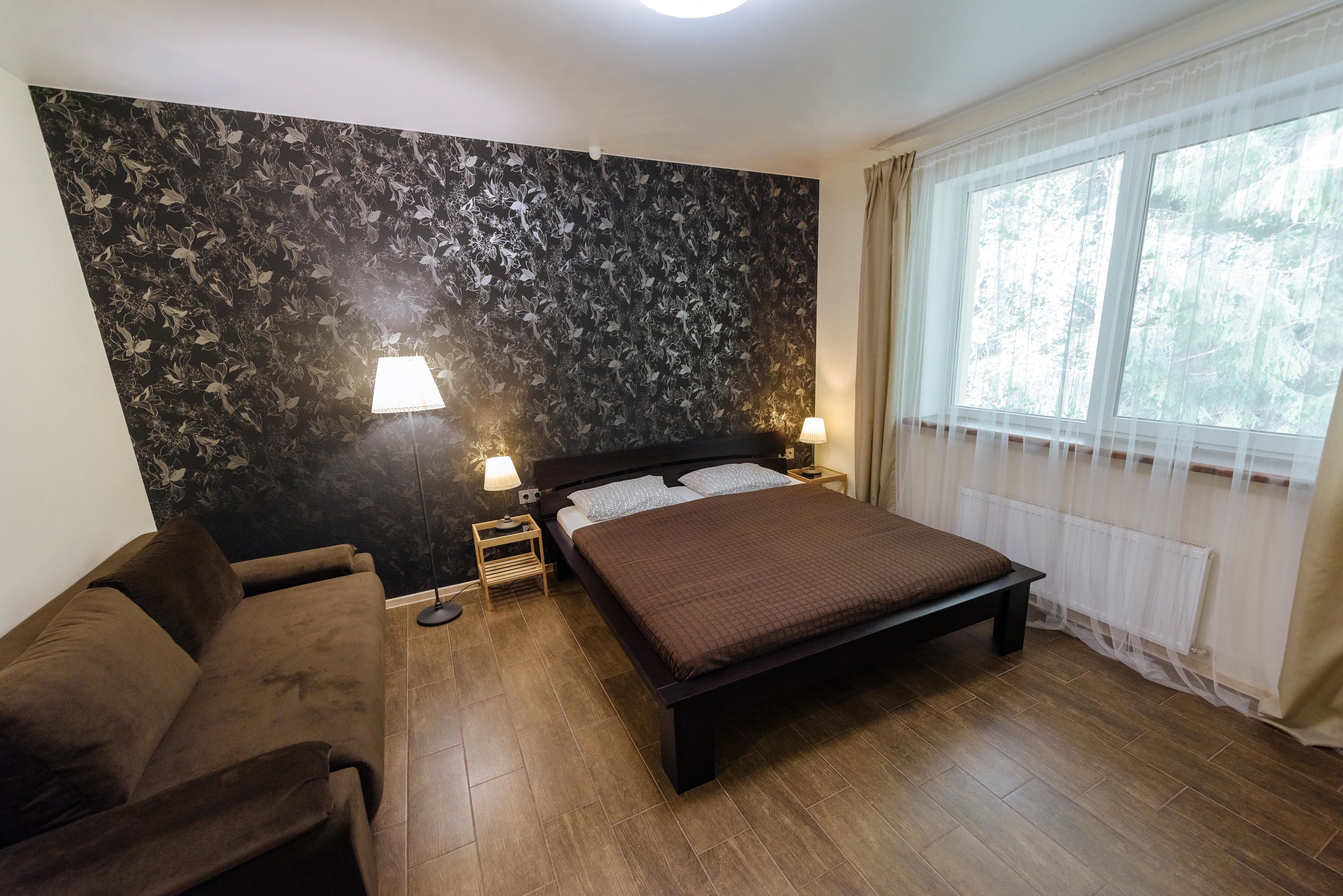 Een slaapkamer met een bed, een slaapbank, twee bedlampjes, een raam met gordijnen en een wand met een bloemenpatroon.