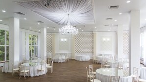 Une pièce spacieuse avec plusieurs tables rondes et des chaises, un grand lustre et de grandes fenêtres ornées de rideaux blancs.