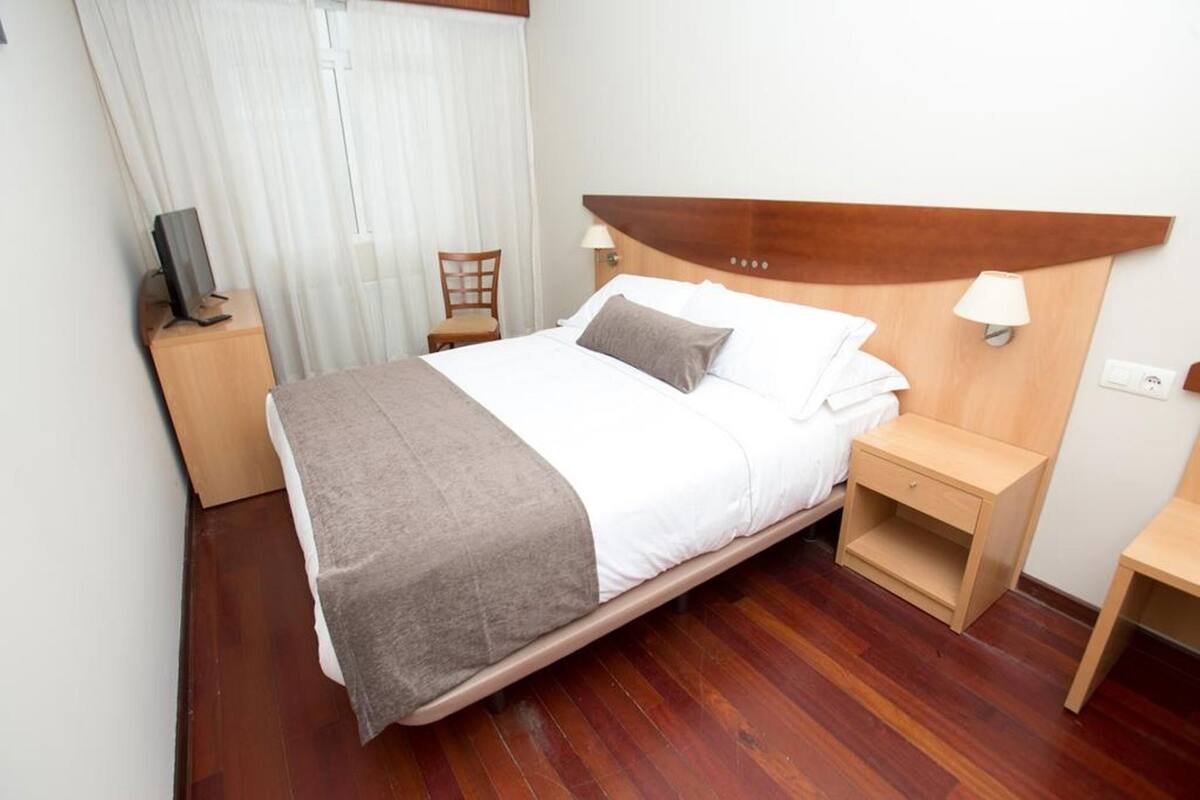 A neatly made bed with a headboard, a bedside table with a lamp, a chair, and a television on a stand.