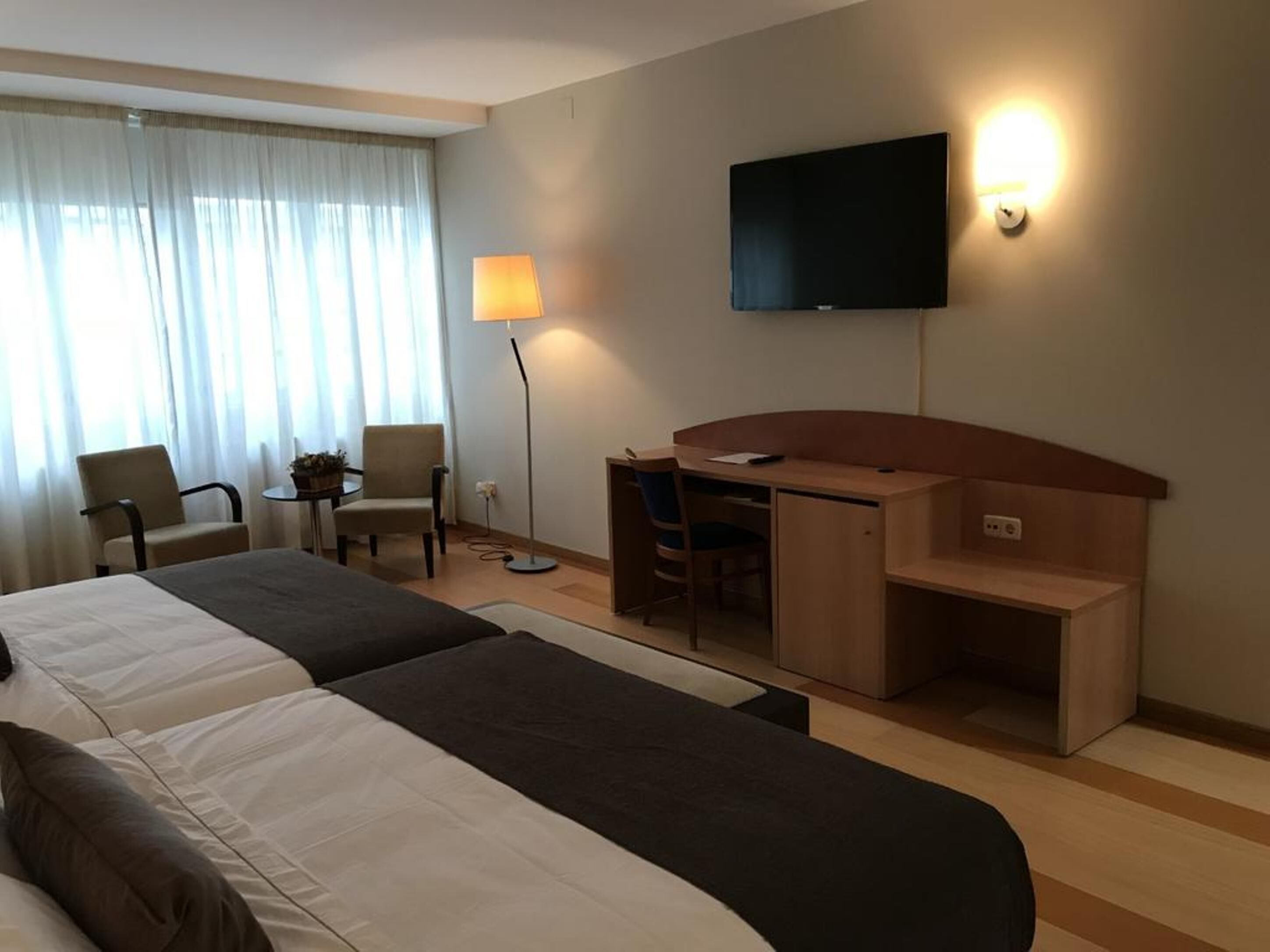A hotel room with two beds, a desk, a chair, a TV, and a lamp.