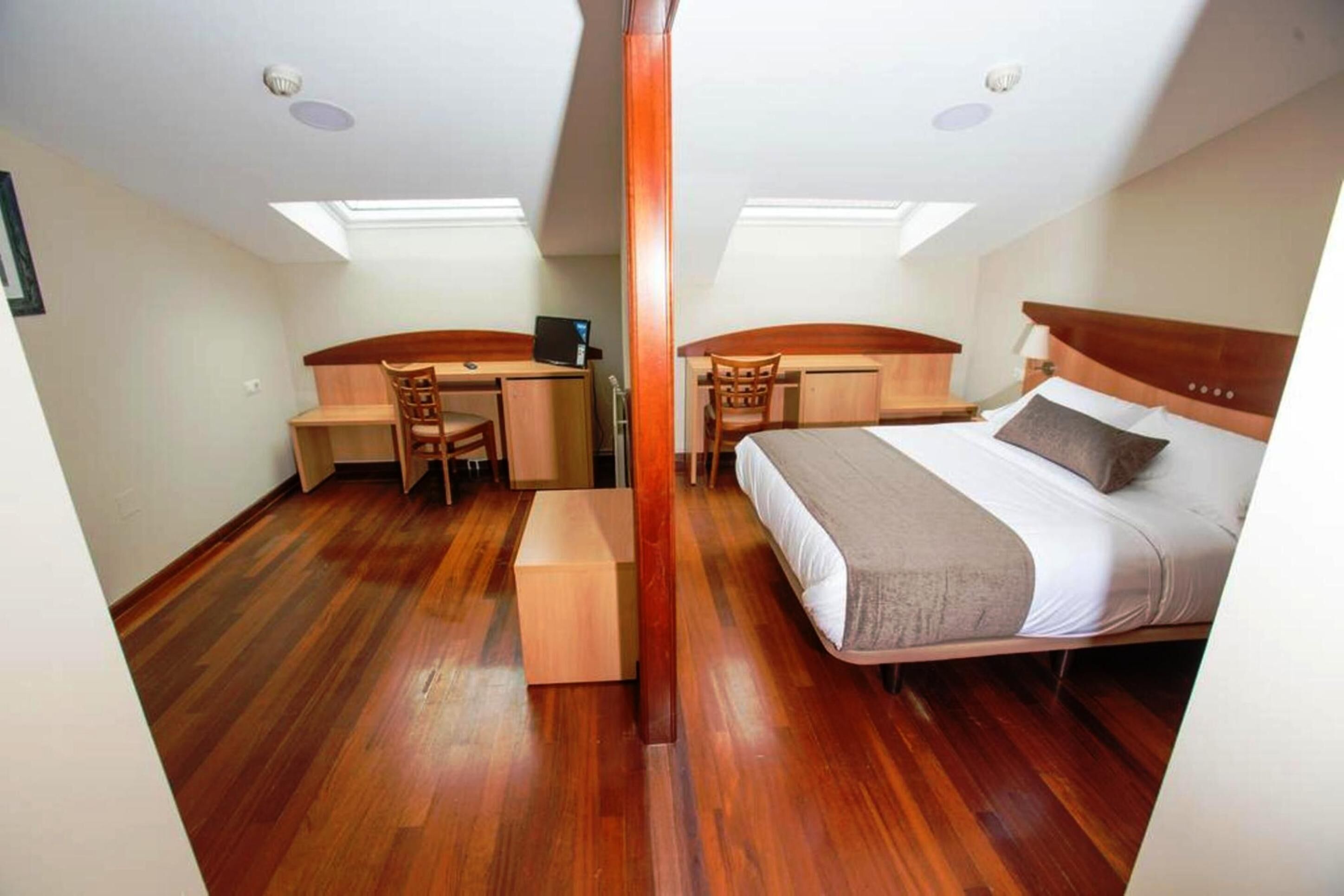 Quarto com cama, escrivaninha e cadeira, com piso de madeira e claraboia.