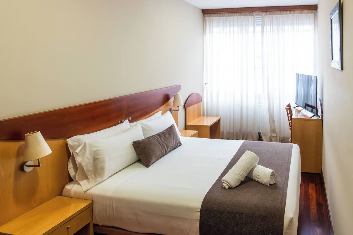 A hotel room with a bed, a nightstand, a lamp, a TV, and a window with curtains.
