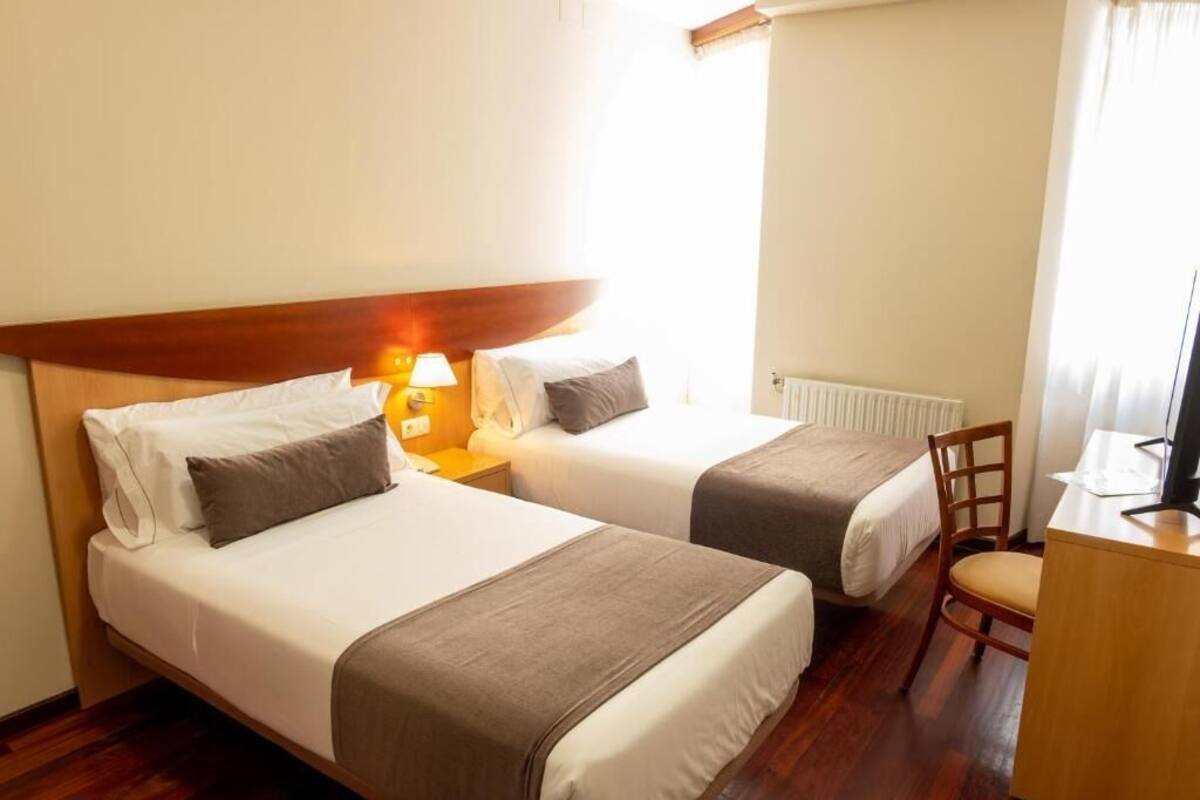 A hotel room with two beds, a desk, a chair, and a lamp.