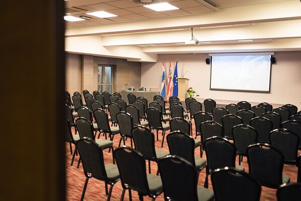 A conference room with rows of black chairs, a podium, and flags on the wall.