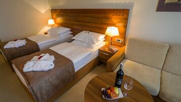 Habitación de hotel con dos camas, un sofá, una mesita de noche con lámpara y una mesa con vino y fruta.