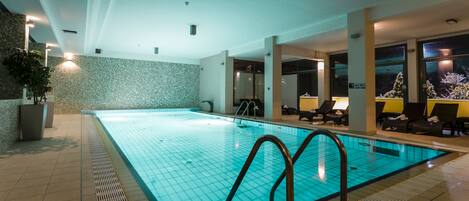 An indoor swimming pool with tiled walls, a tiled floor, and a pool with a grid pattern. There are lounge chairs and a potted plant near the pool.