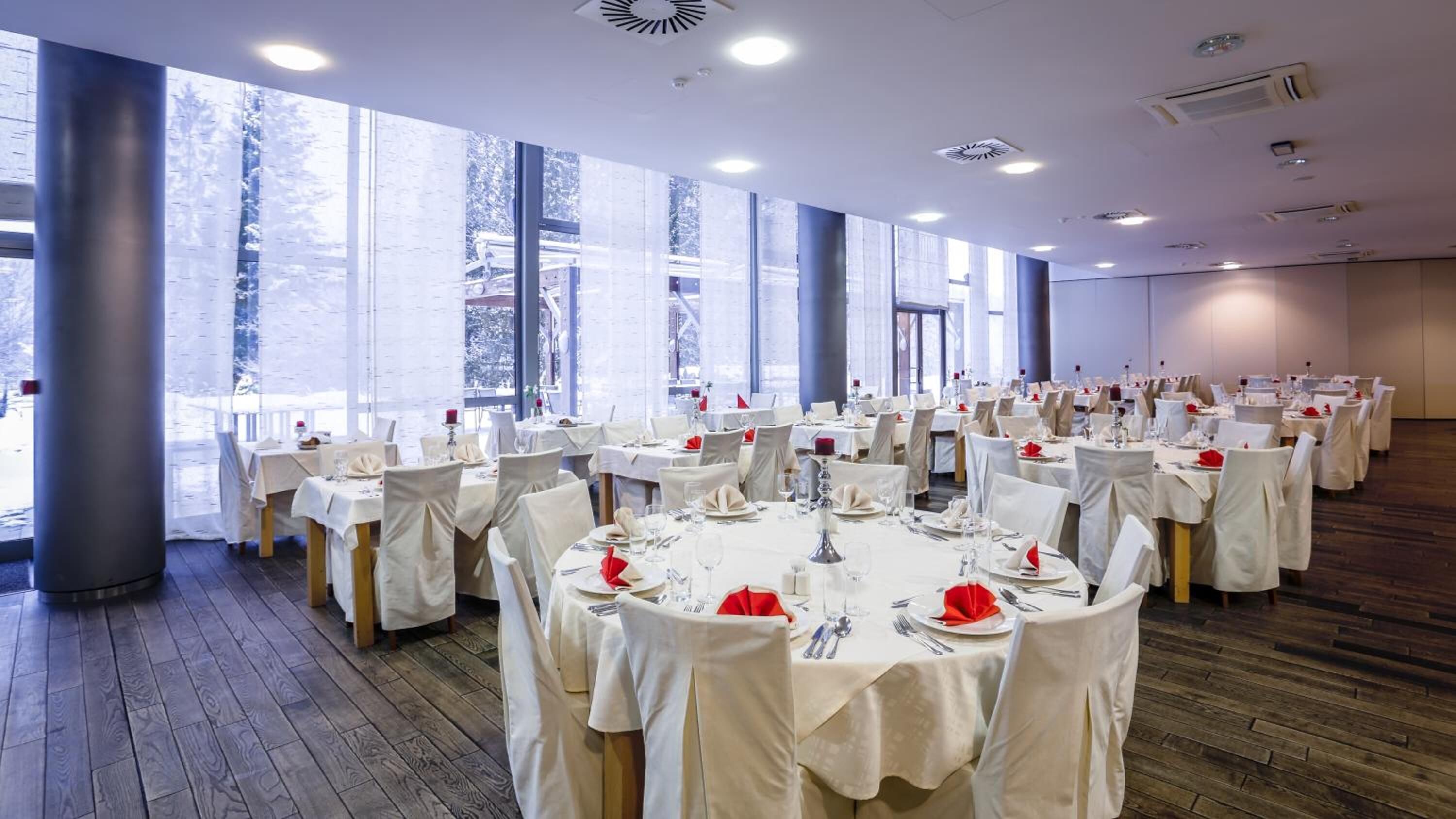 A large banquet hall with round tables set for dining, large windows, and a wooden floor.
