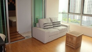 Una sala de estar moderna con un sofá blanco, almohadas a rayas, un baúl de madera y vistas de edificios altos a través de amplias ventanas.